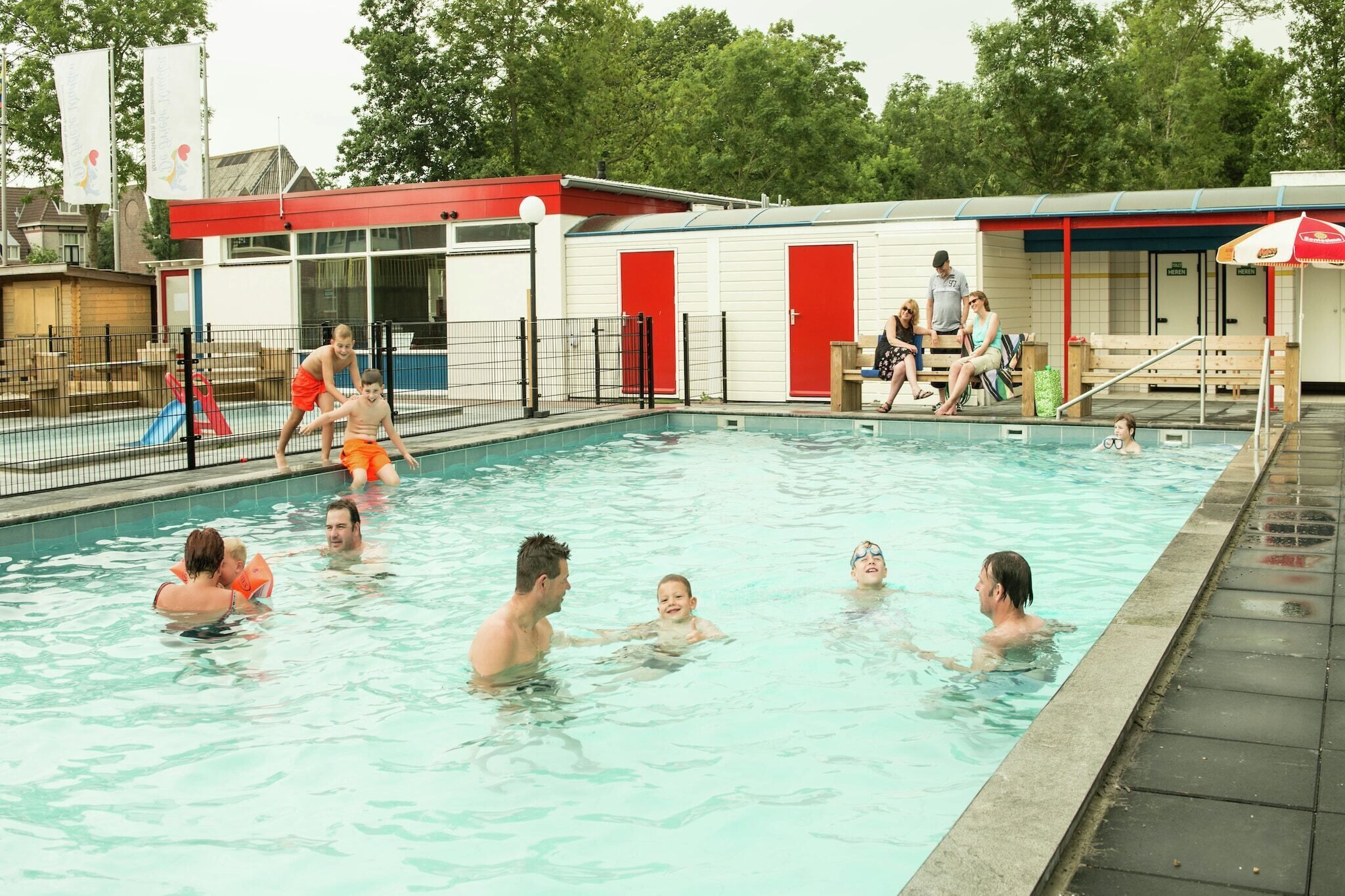 Un espace piscine doté d’une terrasse couverte, de portes rouges et d’une piscine entourée d’une clôture.