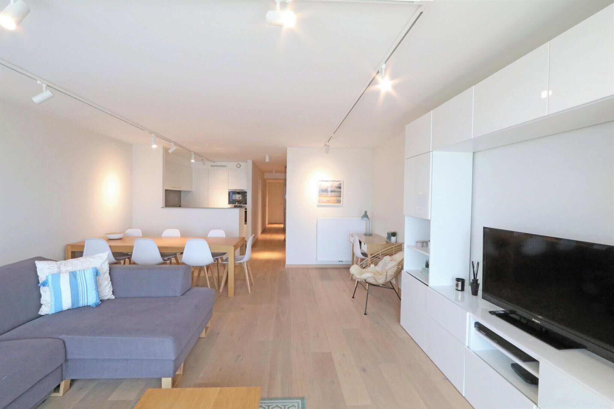 Un salon moderne avec un canapé gris, une table à manger et des chaises, un téléviseur à écran plat fixé sur un meuble blanc, et un coin cuisine à l’arrière-plan.