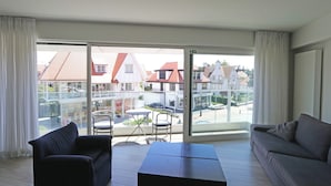 Un balcon avec un canapé et une petite table, une porte vitrée donnant sur la rue et une vue sur les maisons au loin.