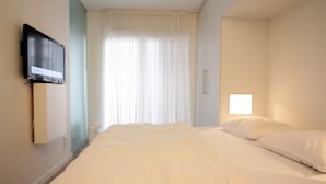 Une chambre d’hôtel avec un lit, un téléviseur fixé au mur, une fenêtre avec des rideaux et une petite table de chevet.