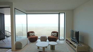 Een moderne woonkamer met een grote schuifpui naar een balkon, een flatscreen televisie en een zithoek met twee fauteuils en een rond zitkussen.
