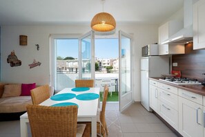 A modern kitchen with white cabinets, a gas stove, and a microwave. There is a dining area with a white table and wicker chairs. - Apartment in Grau du Roi near the Beach (Le Grau-du-Roi)