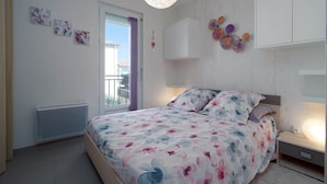 A bedroom with a bed, a bedside table with a lamp, a radiator, and a window with curtains. - Apartment in Grau du Roi near the Beach (Le Grau-du-Roi)