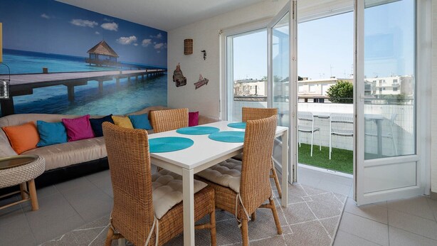 A dining area with a white table and wicker chairs, a sofa with colorful cushions, and a mural of a pier extending into the ocean. - Apartment in Grau du Roi near the Beach (Le Grau-du-Roi)