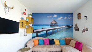 A living room with a large mural of a pier extending into a blue ocean, a beige sectional sofa with colorful cushions, a small round ottoman, and a mounted TV. - Apartment in Grau du Roi near the Beach (Le Grau-du-Roi)