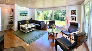 A living room with a sofa, armchair, coffee table, and a view of greenery outside.