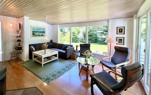 A living room with a sofa, armchair, coffee table, and a view of greenery outside.