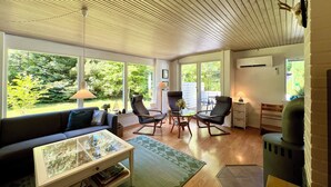 A modern living room with a sofa, coffee table, and chairs, large windows with a view of greenery, and a wood-paneled ceiling.