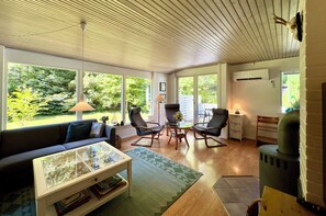 A modern living room with a sofa, coffee table, and chairs, large windows with a view of greenery, and a wood-paneled ceiling.