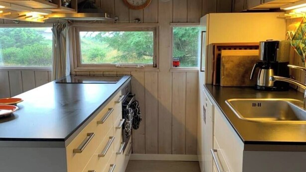 Une cuisine de taille réduite, dotée de meubles en bois, d’une horloge et d’une fenêtre donnant sur un paysage verdoyant.