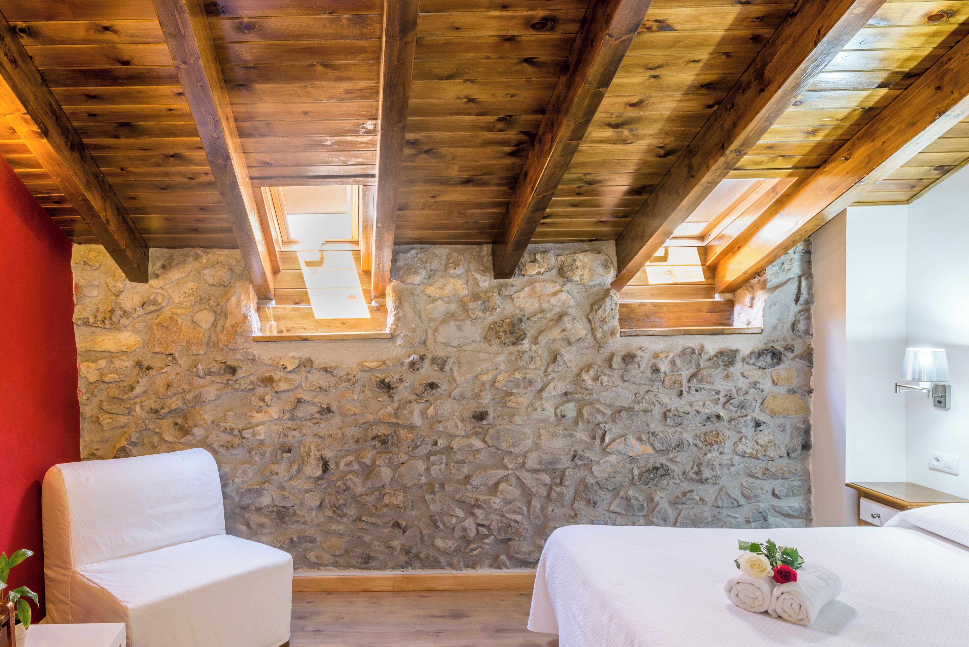 A room with stone walls, wooden beams, a bed with a white bedspread and towels, a chair, and a small table with a plant.