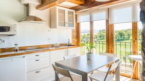 Une cuisine moderne avec des armoires blanches, un four à micro-ondes et un plan de travail en bois. On y trouve une table à manger avec des chaises et une plante en pot. De grandes fenêtres offrent une vue sur un paysage verdoyant.