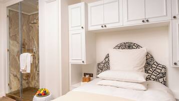 A hotel room with a glass-enclosed shower, a bed with patterned headboard, white cabinets, and a small table with fruit.