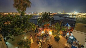 Une terrasse extérieure avec des tables et des chaises, donnant sur un front de mer avec des bâtiments et un pont.