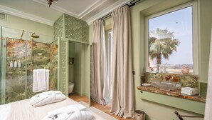 Une chambre d’hôtel avec un grand lit, une douche en verre, un meuble-lavabo avec un vasque et une vue sur les palmiers par la fenêtre.