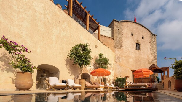 Un espace piscine avec des chaises longues et des parasols, un mur en pierre et une structure en forme de tour.
