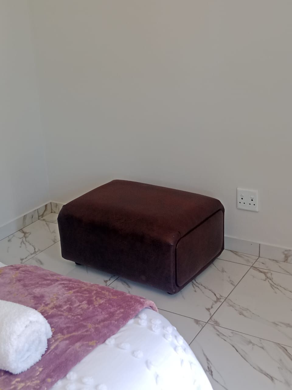 Une pièce comprenant un lit, un pouf marron, une couverture blanche et rose, et un sol carrelé en marbre.