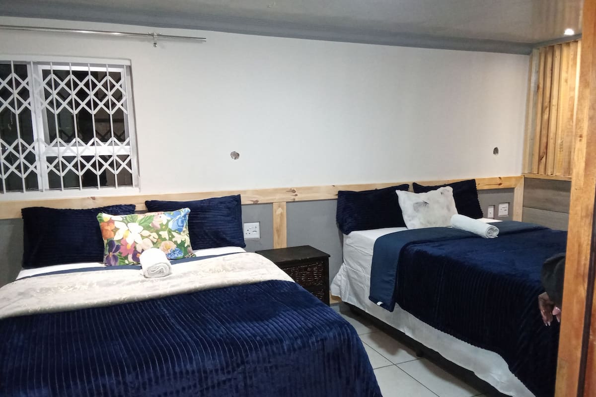 A room with two beds, each with dark blue bedding and decorative pillows. There is a window with metal bars and a wooden headboard.