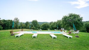 Une piscine rectangulaire bordée de chaises longues blanches et ombragée par des parasols verts, située dans un espace verdoyant entouré d’arbres.