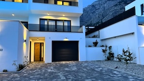 A modern multi-story building with a central garage door, a balcony on the upper floor, and a landscaped area with small trees. - NEW Villa Dado with Pool, sea view (Makarska)