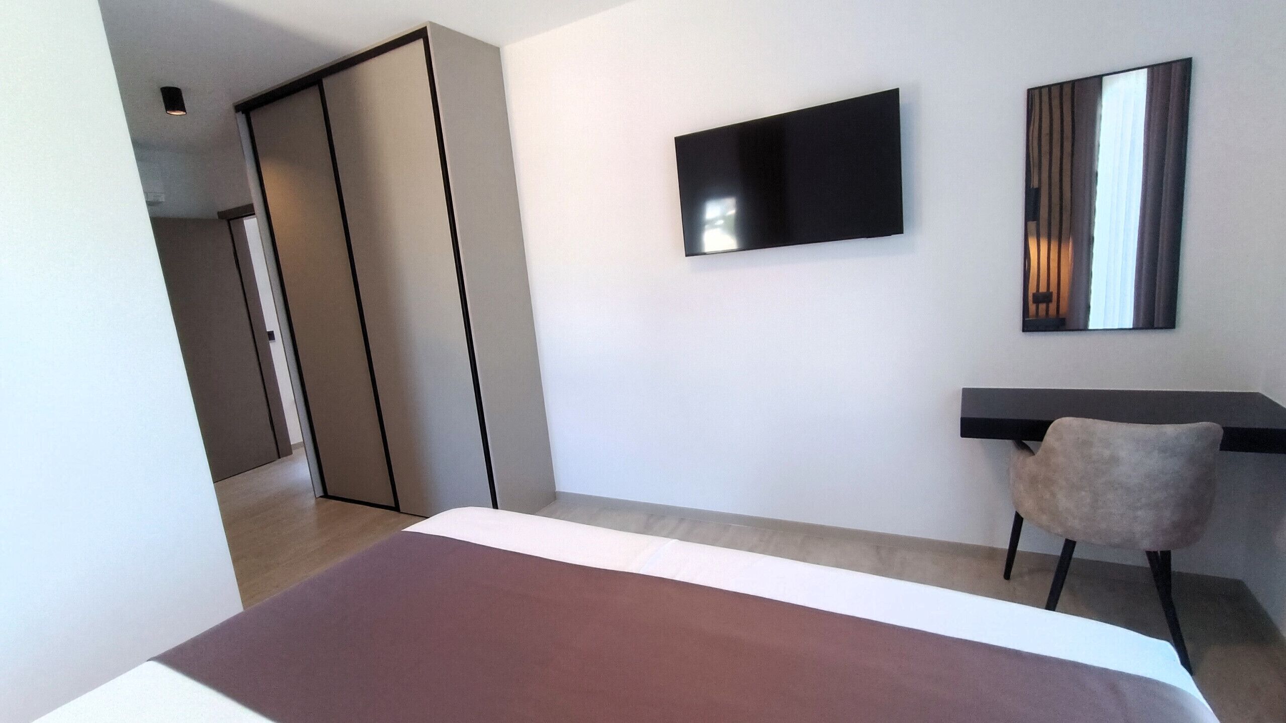 A modern hotel room with a bed, a desk, a chair, a TV, and a wardrobe.