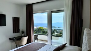 A hotel room with a bed, a desk with a chair, a TV, and a balcony with a view of the sea and buildings.