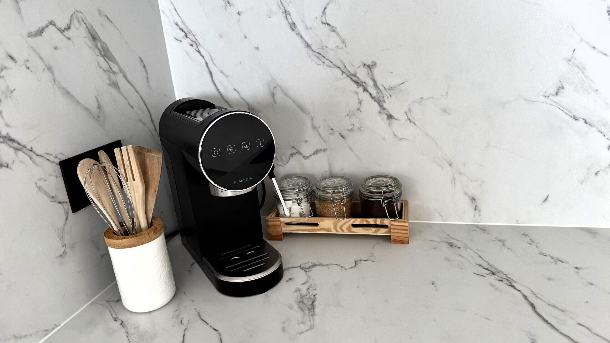 A kitchen with a coffee maker, utensils, and jars on a marble countertop.