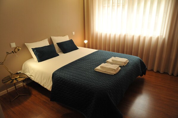 A neatly made bed with a dark quilt, two pillows, and folded towels on a wooden floor.