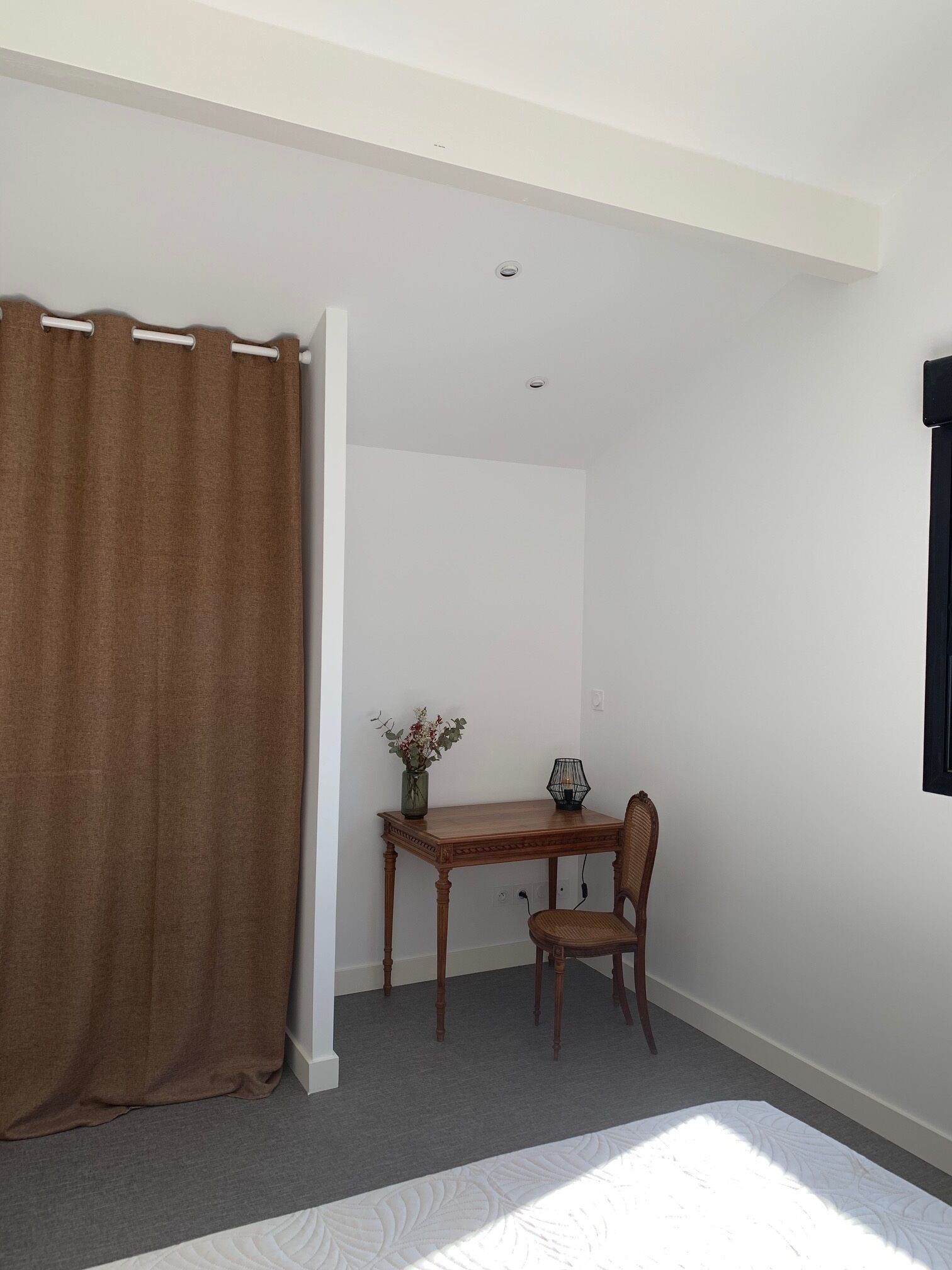 A room with a bed, a wooden table with a chair, a vase with flowers, and a black lamp.
