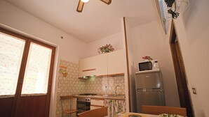 Eine kleine Küche mit Kühlschrank, Mikrowelle und Herd. Ein Esstisch mit Stühlen und ein Fenster mit blumigen Vorhängen.