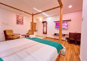 Quarto de hotel com uma cama grande, duas cadeiras, uma mesinha, uma televisão e estrutura de teto em madeira.