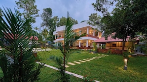 A modern building with a flat roof and multiple balconies, surrounded by well-maintained greenery and a paved pathway. - Voye Homes Signature Pool Resort Varkala (Varkala)