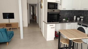 A modern kitchen with white cabinets, a microwave, oven, and dishwasher. There is a wooden table with two stools, a TV, and a blue sofa in the living area. - Bright cocoon with private courtyard (Salses-le-Château)
