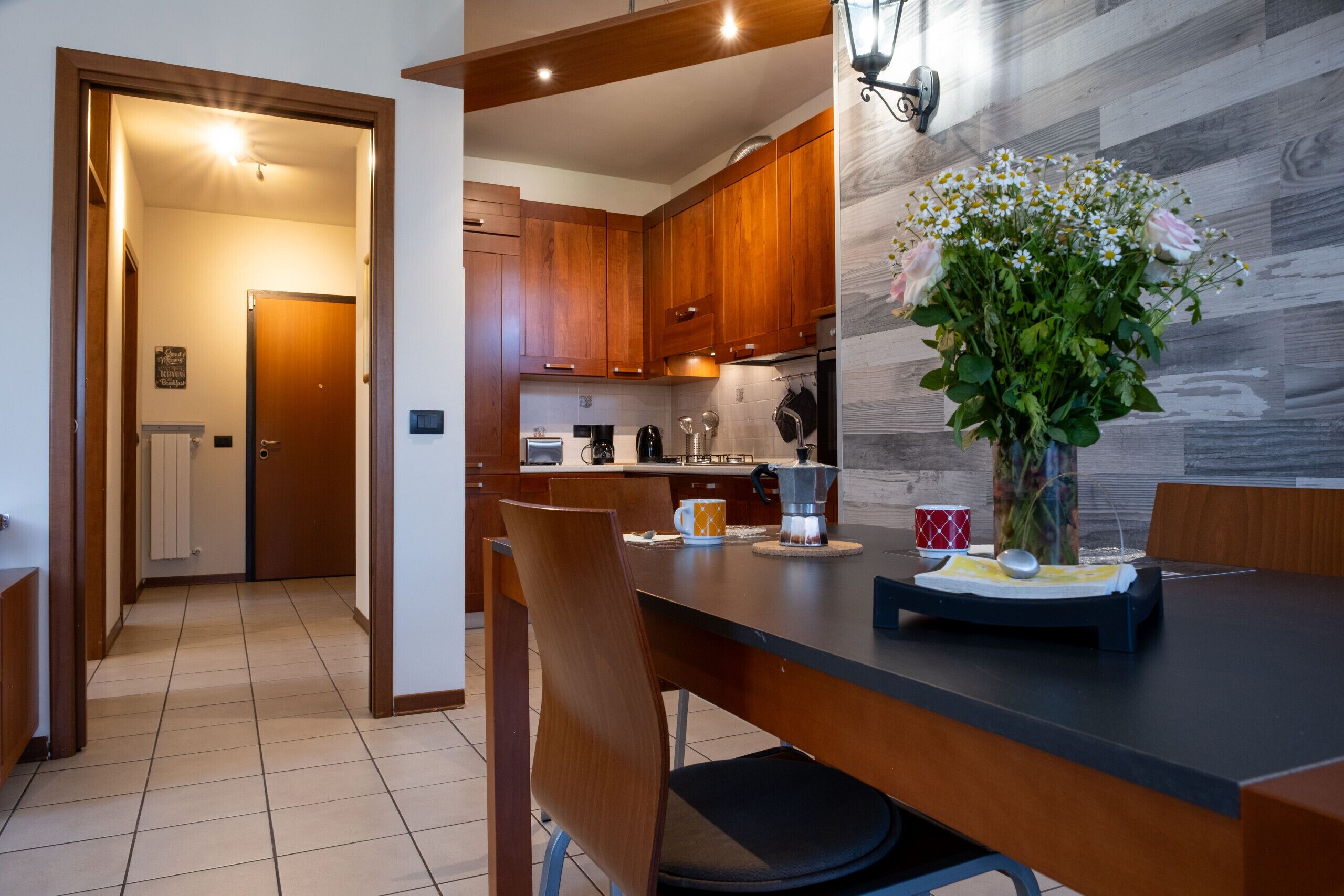 A modern kitchen with wooden cabinets, a dining table with chairs, and a vase of flowers on the table.