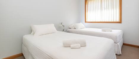 A room with two single beds, each with a pillow and a rolled towel, a bedside table with a lamp, and a window with blinds.