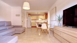 A modern living room with a grey sofa, a dining area with a table and chairs, a kitchen in the background, and a TV on a wooden cabinet. - Anna del Mar | Coastal Boho (San Luis de Sabinillas)