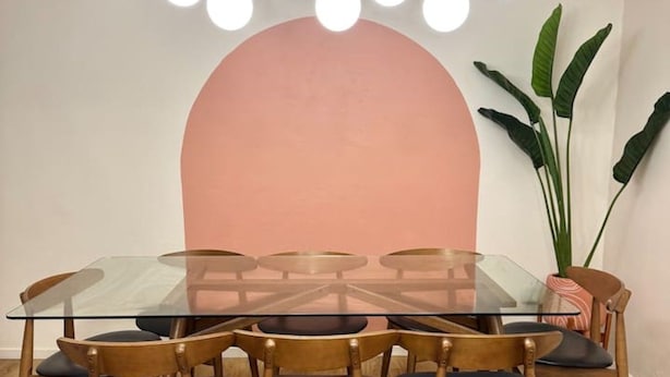 Un comedor con una mesa de vidrio rodeada de sillas de madera, un arco de pared rosa y una lámpara colgante con múltiples bombillas.