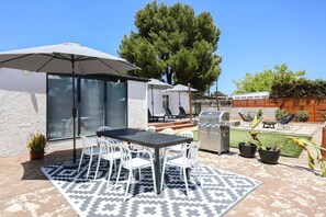 Een terras buiten met een tafel, stoelen, een barbecue en een grote parasol.
