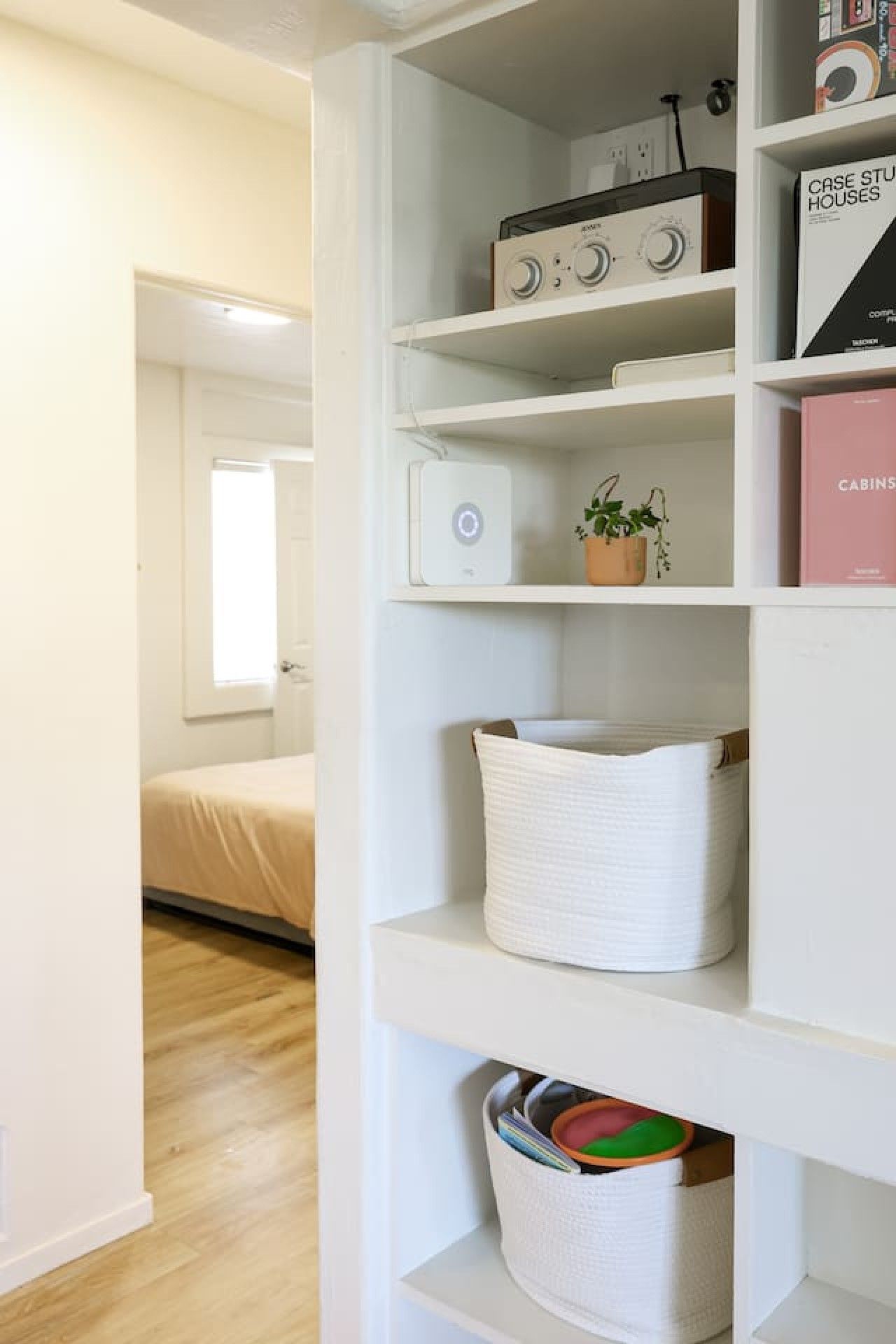 A white interior with a bed, a white shelf unit containing a radio, books, and a plant.