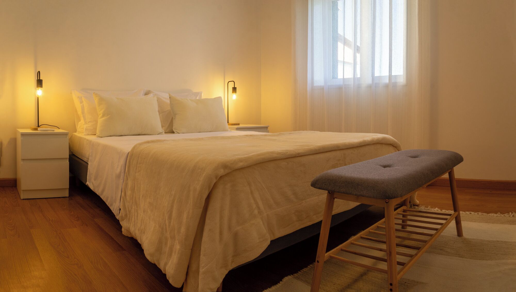 Un dormitorio ordenado con cama, mesitas de noche, un banco y una ventana con cortinas traslúcidas.