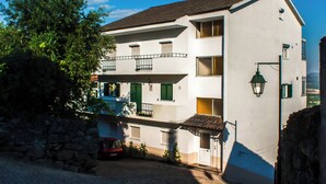 Un immeuble de deux étages doté d’un balcon, d’un mur en pierre et d’une voiture rouge garée devant.