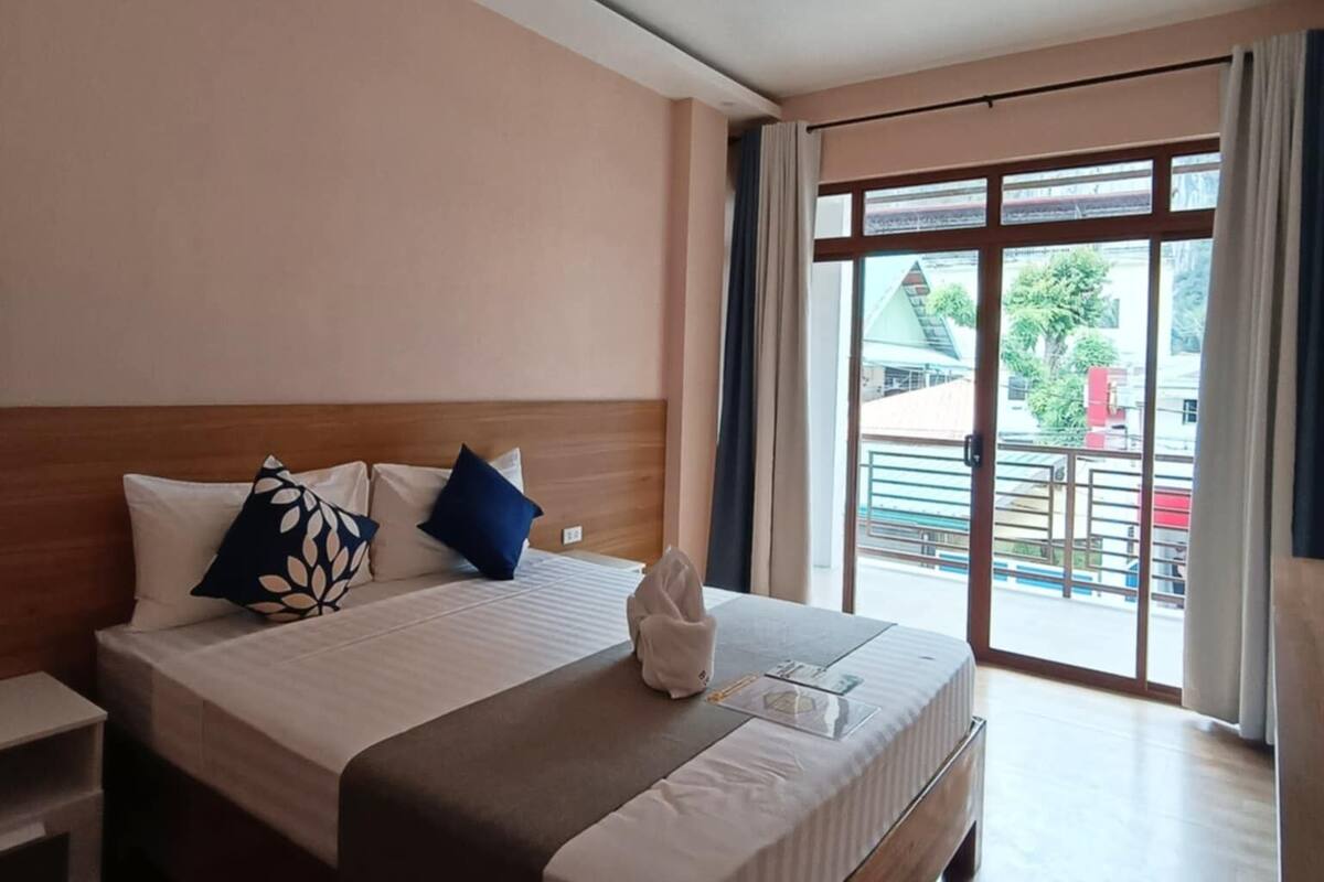 A hotel room with a large bed, a wooden headboard, a bedside table, and a view of the outside through a sliding glass door.