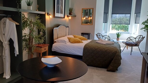 Een moderne slaapkamer met een bed, nachtkastje en een rond tafeltje met een kom. Er staan potplanten en er hangt een spiegel aan de muur.