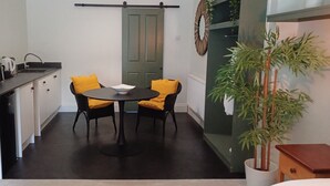 Een compacte keuken met eethoek, bestaande uit een zwarte tafel, twee stoelen met gele kussens, een potplant en een keukenblad met een waterkoker.