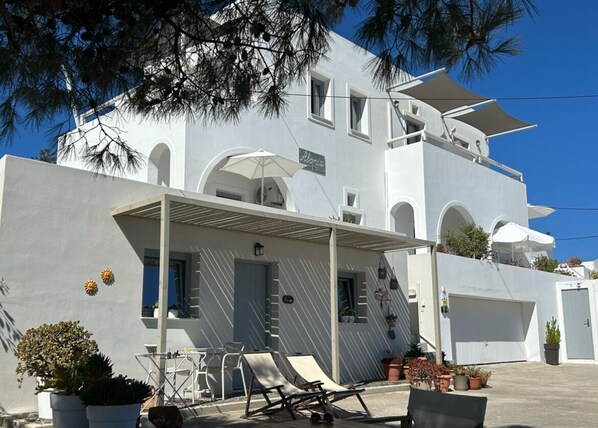 A modern white building with a covered patio, outdoor seating, and potted plants. - Alonia Studios (Santorini)