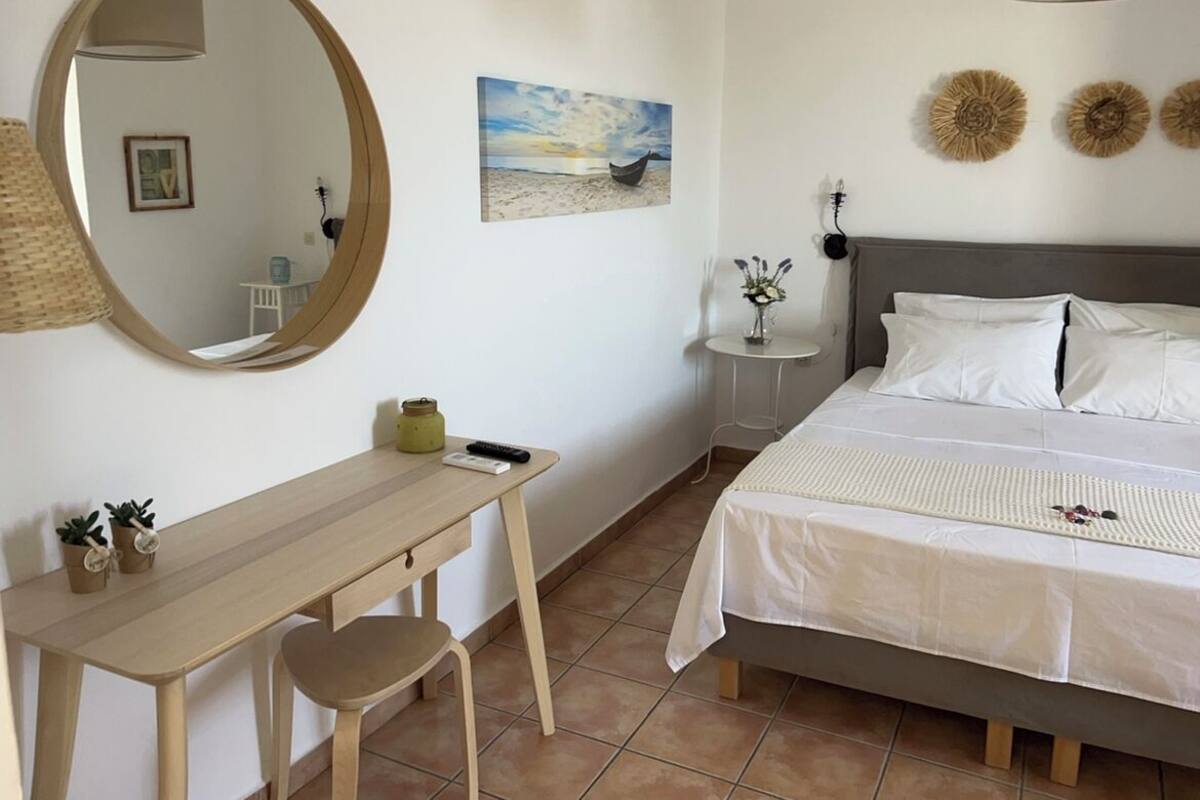 A bedroom with a bed, a desk, a chair, a mirror, a small table with a vase, and a painting on the wall.