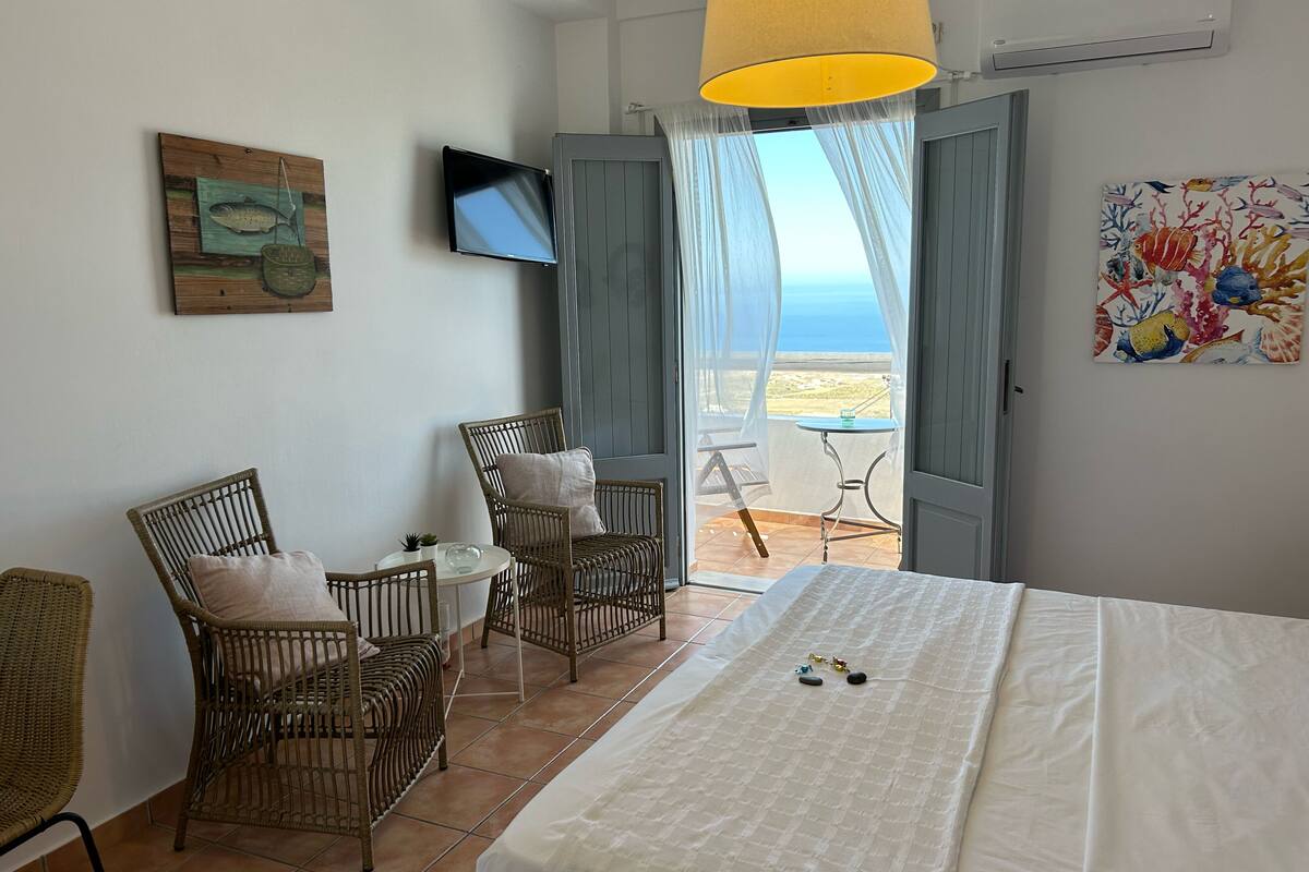 A bedroom with a bed, two wicker chairs, a small table, a TV, and a view of the sea through a window.