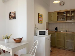 A compact kitchen with a microwave on a small fridge, a sink, wooden cabinets, and a wall clock. - Alonia Studios (Santorini)