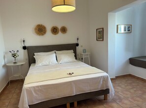 A neatly made bed with white bedding, a bedside table with a vase of flowers, and decorative wall art. - Alonia Studios (Santorini)