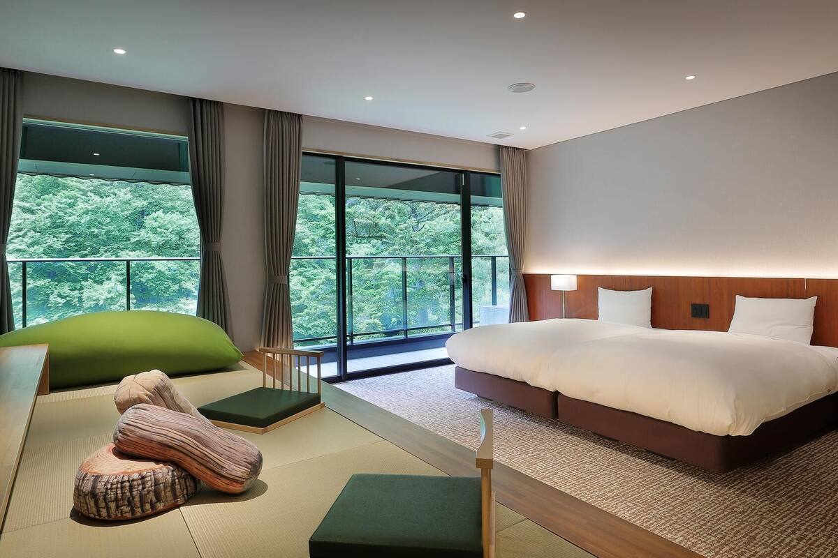 A modern hotel room with a large bed, a green sofa, a wooden side table, and a view of lush greenery through large windows.